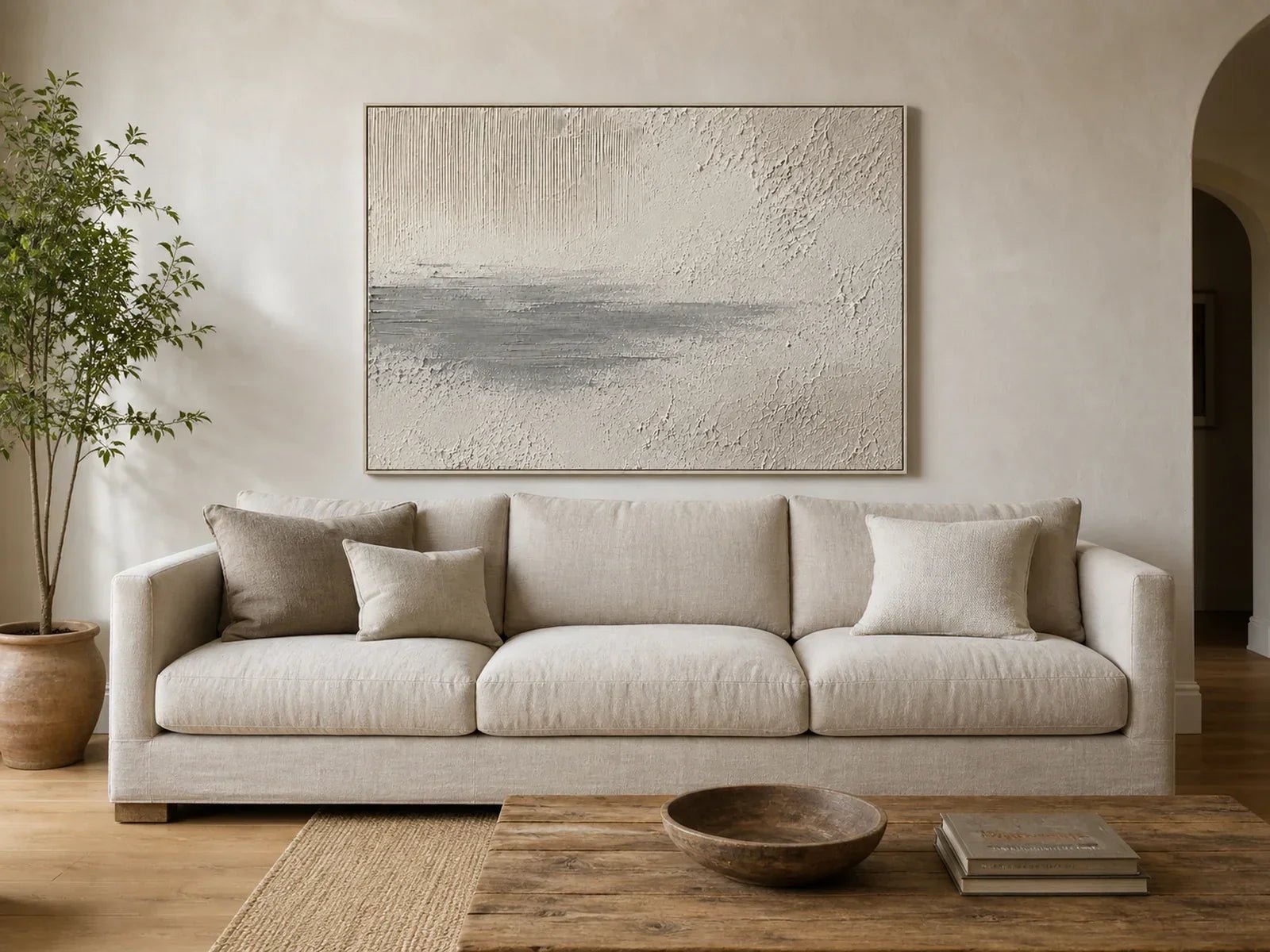Wide neutral textured artwork centered above an oatmeal three-seat sofa in a naturally lit living room.
