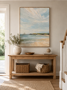 Neutral coastal landscape painting displayed above a wood console in a modern organic entryway