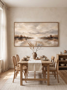 Framed horizontal soft taupe river landscape painting displayed in open-concept dining room with wood furniture