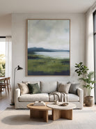 Vertical green landscape canvas painting with lake view mounted above a modern neutral living room sofa