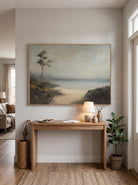Framed peaceful coastal landscape painting styled above a wooden console table in a bright entryway